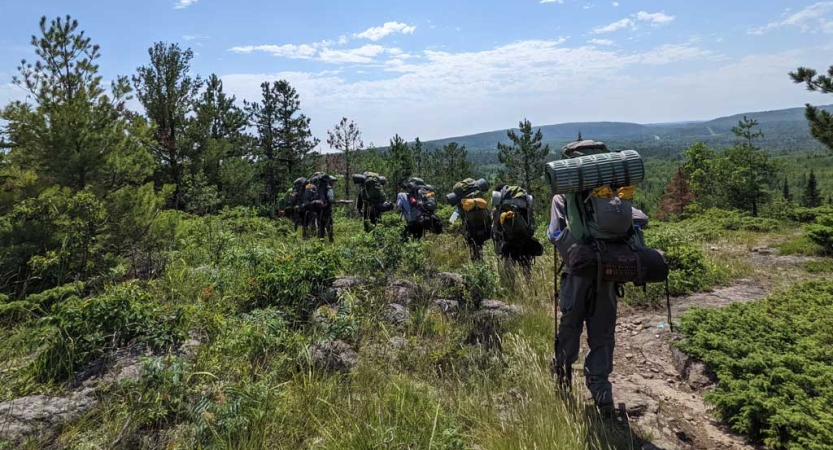 A group of students carrying backpacks hike along a trail in an open green area. They are walking towards trees, and there are green hills in the background. 